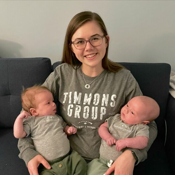 Hannah wearing a gray Timmons Group shirt holding her twin babies who are wearing matching gray Timmons Group onesies.