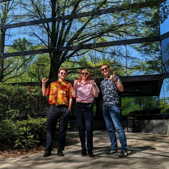 Two men and a woman wearing Hawaiian shirts and matching sunglasses while posing with their pinky and thumbs out on their hands.