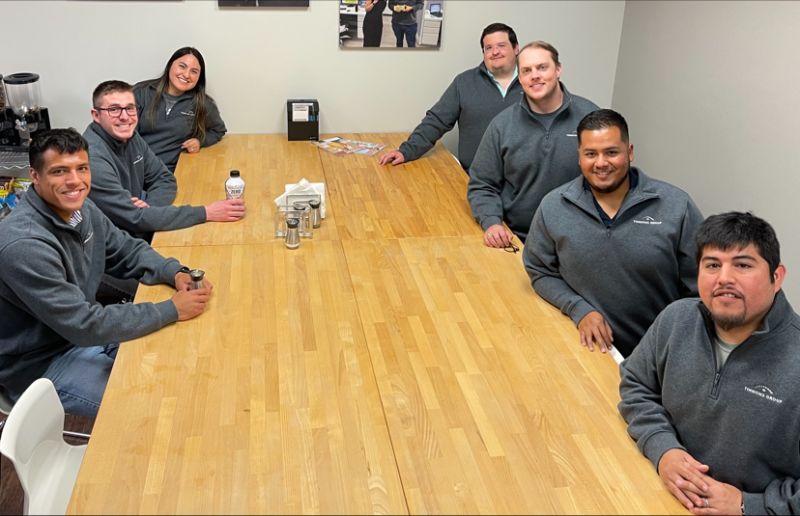 Six men and one woman all sitting at a conference table while wearing gray quarter zip Timmons Group pullovers.