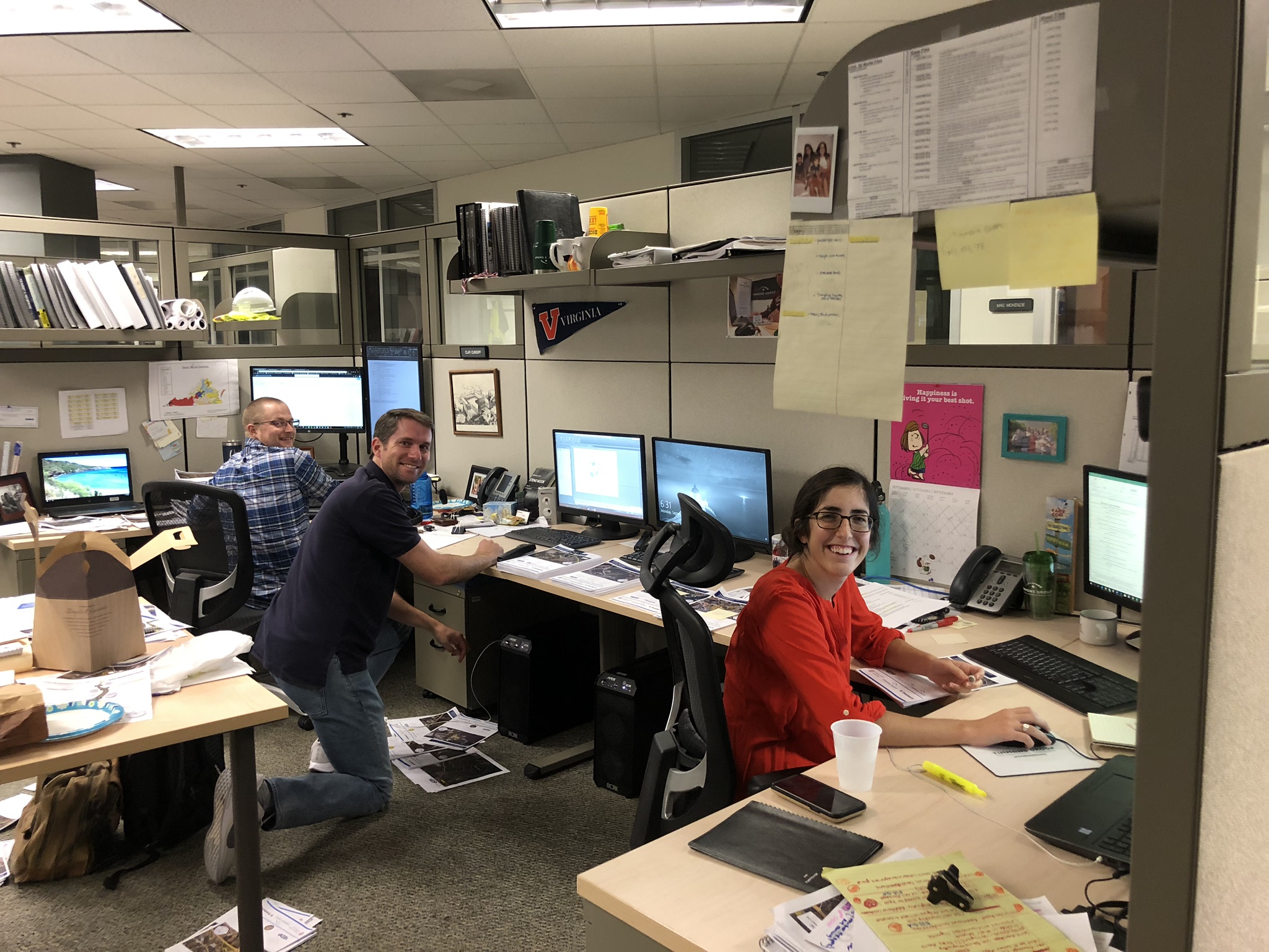 Leandra at her desk with two male coworkers behind her posing for picture.