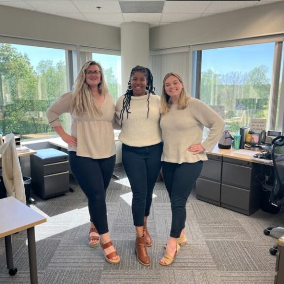 Three women wearing white shirts, blue denim jeans, and brown shoes.