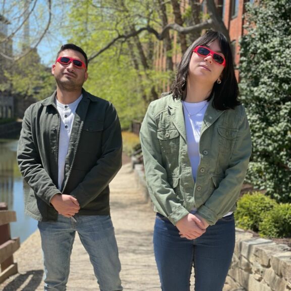 George on the left and Savanna on the right wearing matching red sunglasses, green jacket, white shirt, and jeans while making the same pose.