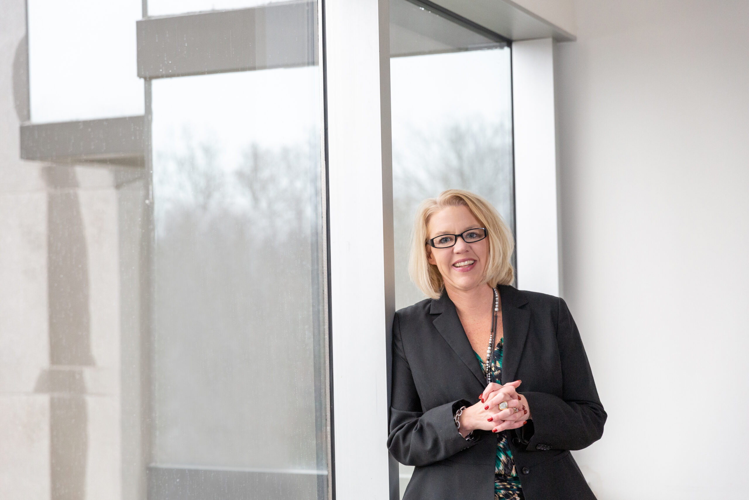 Shannon wearing a black blazer, leaning against a wall with her hands clasped together.