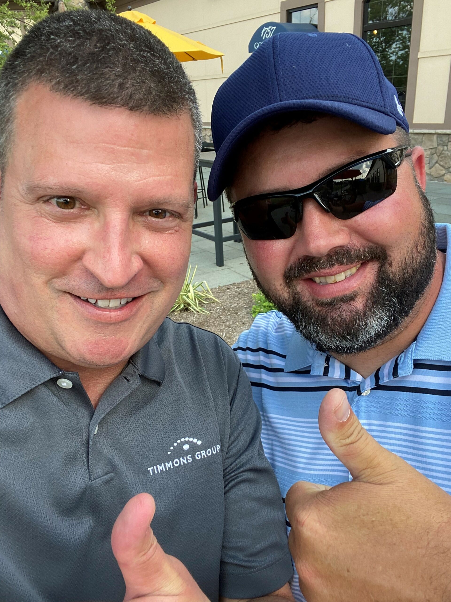 Mike Newman (left) wears a grey Timmons Group polos shirt. Justin Lilly (right) wears a light blue polo shirt, navy blue ball cap, and sun glasses. They are giving thumbs up.