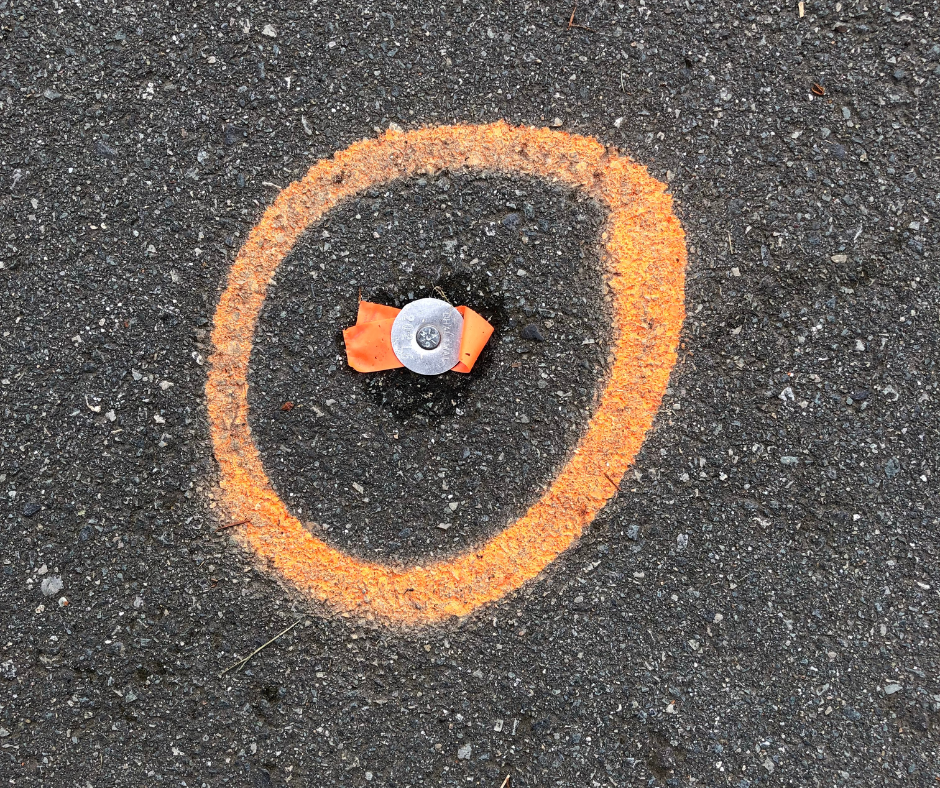 A marker on the road is circled with orange spray paint and tied with orange survey tape.