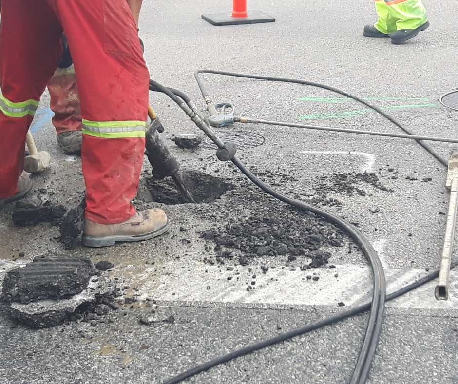 A worker wearing reflective red and yellow work pants drills into an asphalt road to identify an underground utility.