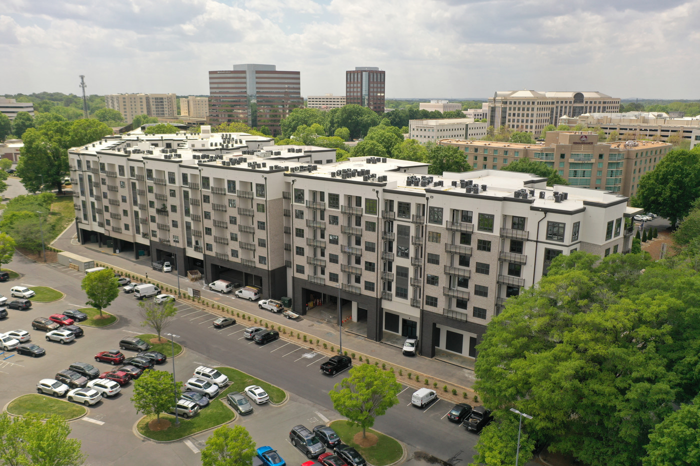 Photo of the outside of Hazel SouthPark showcasing the 6-story apartment building and its balconies. The building is beige, cream, and gray colored. The background features the city of Charlotte. Parking is available outside of the building.