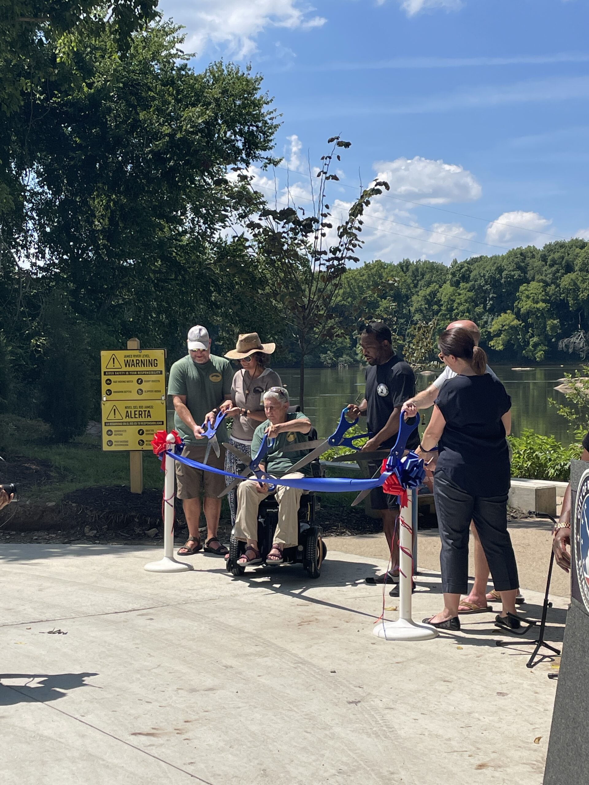 A picture of Richmond Mayor Levar Stoney and members of the James River Outdoor Coalition cutting the blue ribbon with giant scissors. They are facing the crowd with the ramp behind them.