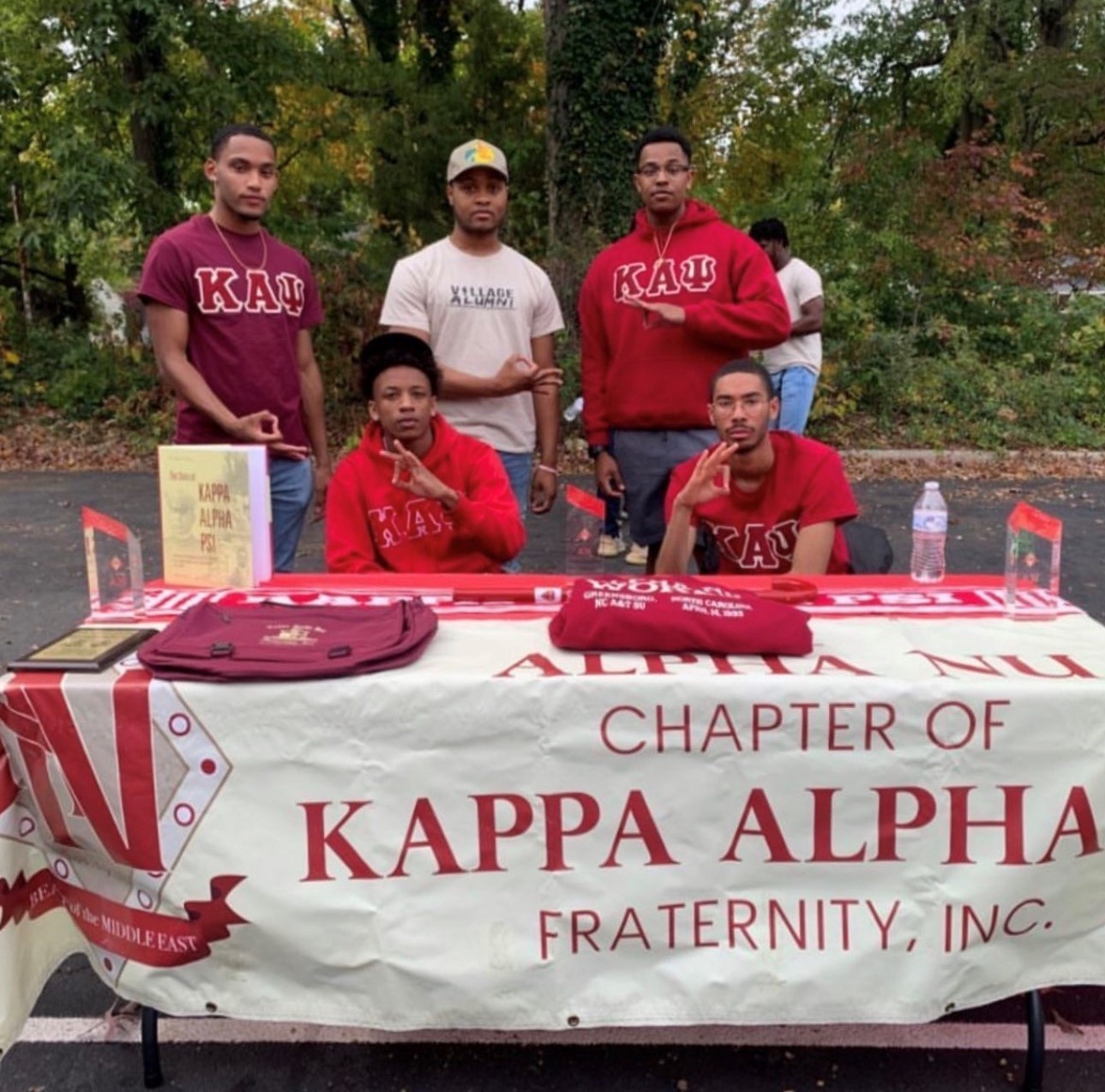 Jalen with his brothers from Kappa Alpha Psi Fraternity, Inc. They are wearing their fraternity letters on red shirts and hoodies while posing for a photo in front of a registration table with a Kappa Alpha Psi table banner that reads "Alpha Nu Chapter of Kappa Alpha Fraternity, Inc." Some of the words were cut from the picture.