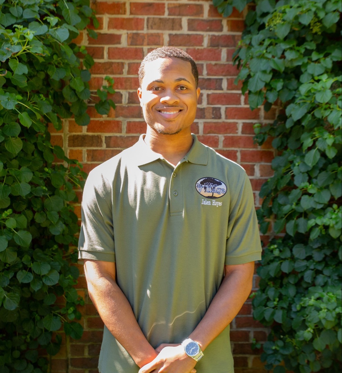 A picture of Jalen Hayes wearing a sage green shirt with The Village Mentoring program logo on the right side. The logo features a tree behind the words "The Village." He is standing in front of a brick wall with ivy on either side of him.