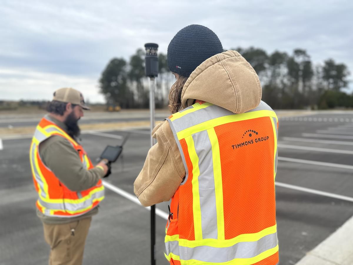 Two of Ed's team members are collecting data points in a parking lot. They are wearing bright orange and yellow PPE vests.