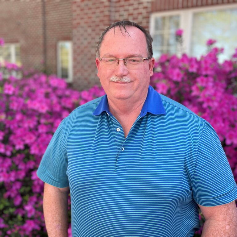 Photo of Ed Jordal wearing a striped blue shirt standing in front of Azalea shrubs that have bloomed with pink Azalea flowers.