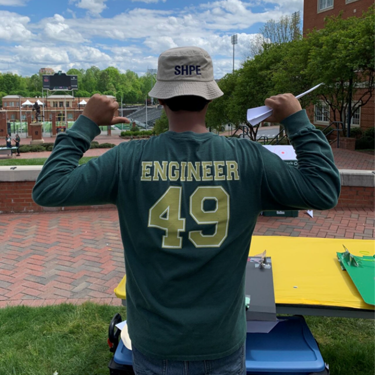 A photo of Erik on the UNC Charlotte campus standing in front of a yellow table. He's facing away from the camera using his thumbs to point at the back of his shirt which reads "Engineer 49." He's also wearing a tan bucket hat that has SHPE on it.