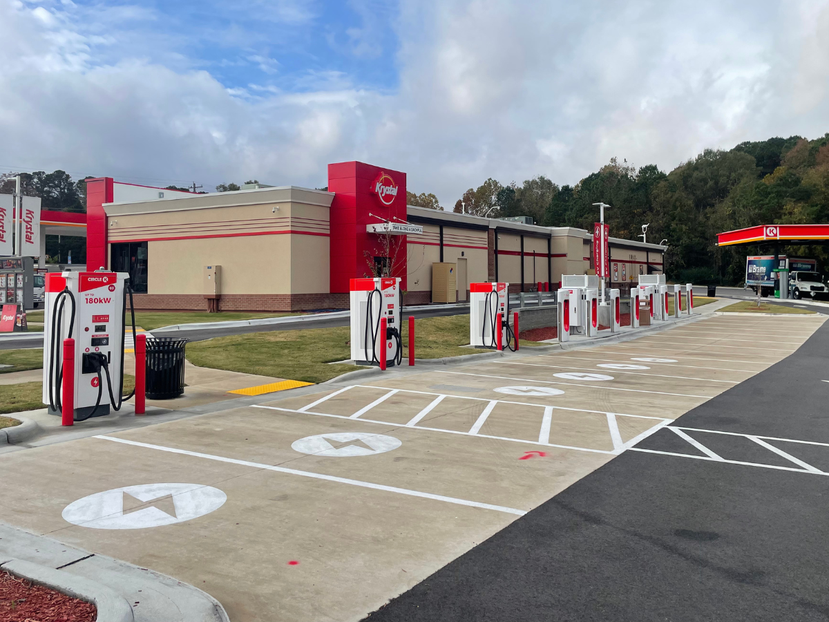 A row of white and red electric vehicle charging stations at the back of a Circle K. Painted in the parking spots are white circles with lighting bolts in the middle.