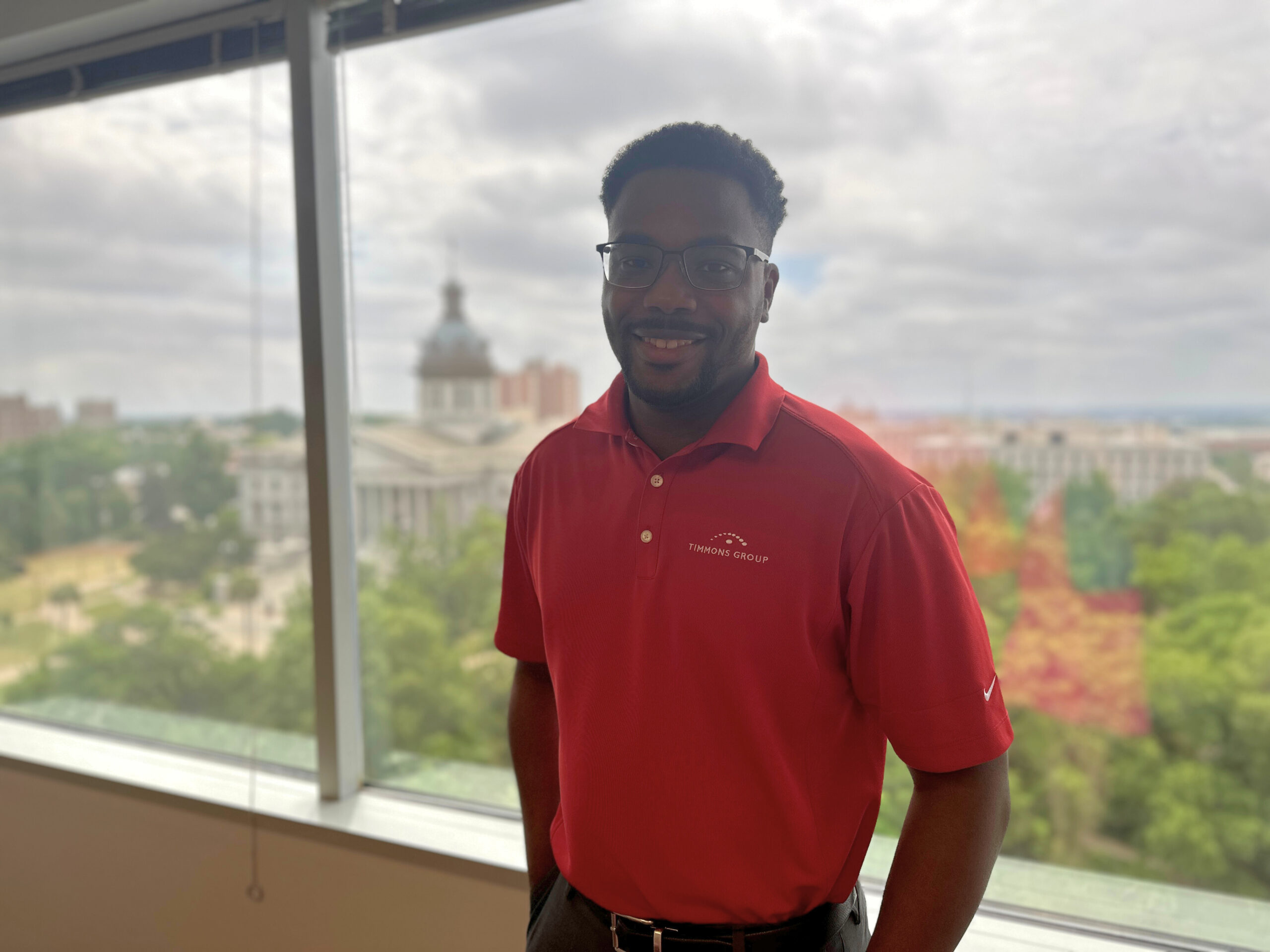 Cliff Lawson standing in front of the office window smiling for a photo with the South Carolina State House in view behind him.