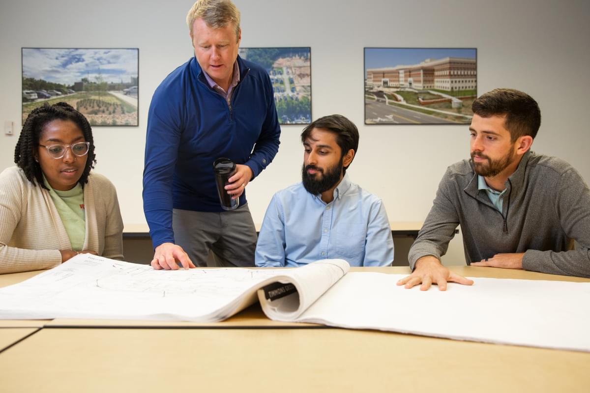 Four Timmons Group employees pictured in a row with three sitting at a table and one standing over the table looking down at plans.