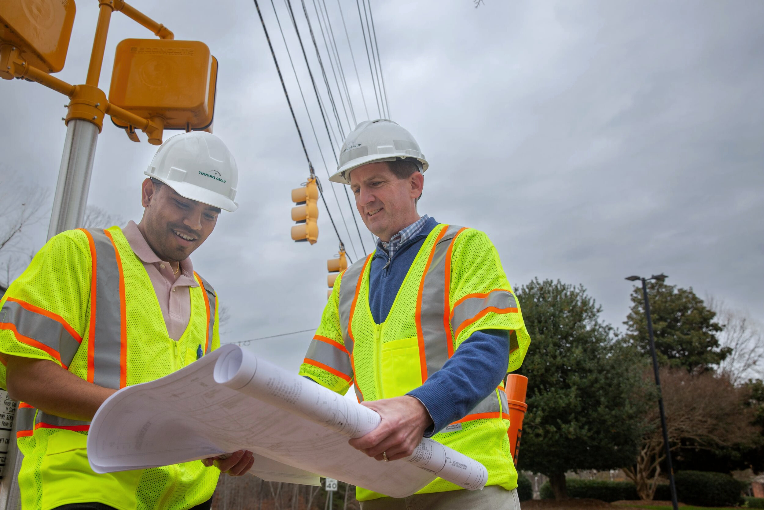 Two Timmons Group traffic engineers pictured wearing high visibility safety vests and white hard hats, standing underneath a traffic light and cross walk signal while holding and looking down at plans.