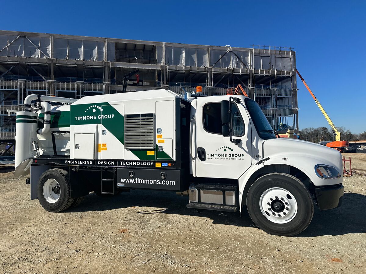 Timmons Group's white and green Subsurface Utility Engineering (SUE) vacuum excavator truck parked in front of a building under construction. The truck has the Timmons Group logo with the "Engineering, Design, Technology" tagline as well as the website www.timmons.com.