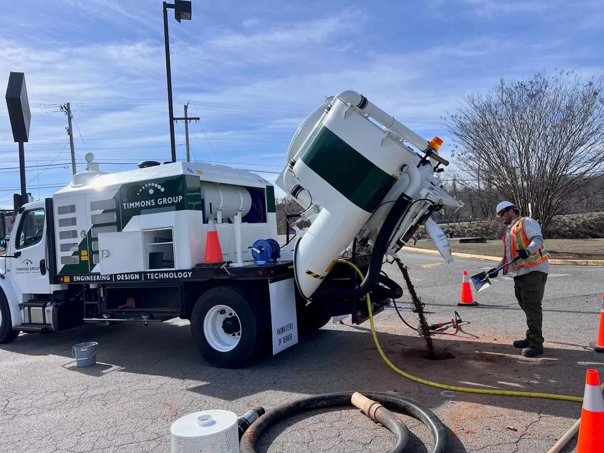 An employee is featured operating the white and green Subsurface Utility Engineering (SUE) vacuum excavator truck while wearing personal protective equipment. The truck has the Timmons Group logo with the "Engineering, Design, Technology" tagline as well as the website www.timmons.com.