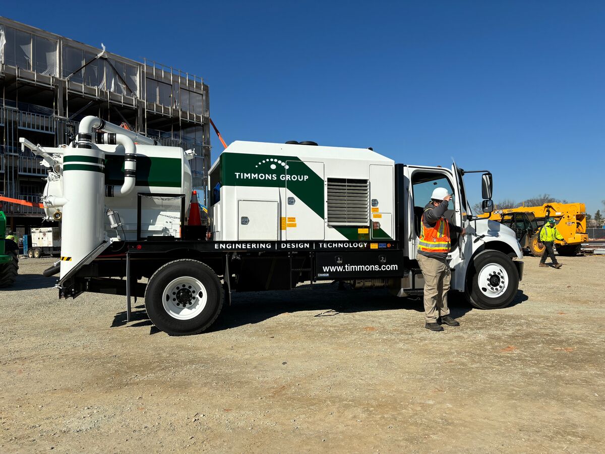 Timmons Group's white and green Subsurface Utility Engineering (SUE) vacuum excavator truck parked in front of a building under construction featuring an employee wearing personal protective equipment (PPE) while stepping out of the truck. The truck has the Timmons Group logo with the "Engineering, Design, Technology" tagline as well as the website www.timmons.com.